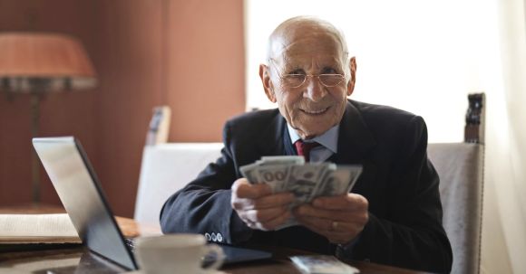 Retirement Management. - Confident senior businessman holding money in hands while sitting at table near laptop